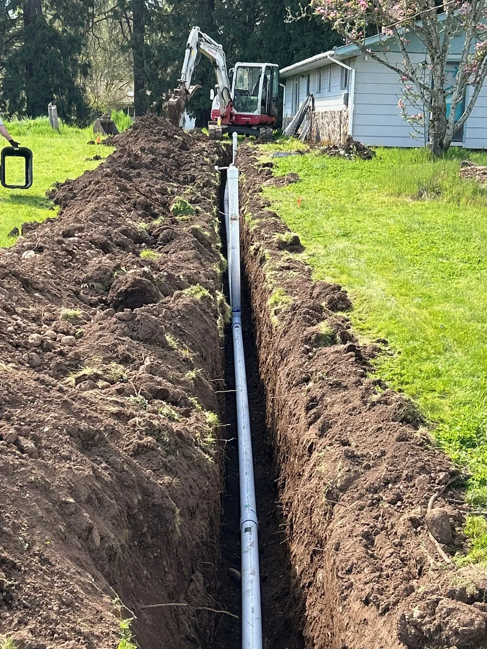 White pipe laid in a trench beside a home with an excavator at the far end